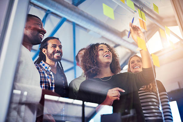 marketing digital b2B un groupe de collègues souriants collaborant activement. Une femme est en train de placer ou d'écrire sur un post-it sur un tableau en verre, utilisé pour le brainstorming ou la gestion de projet. L'équipe affiche une dynamique positive et une ambiance de travail coopératif.