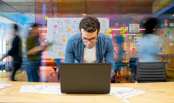 marketing digital b2B. Un homme concentré travaillant sur un ordinateur portable dans un bureau animé. Derrière lui, un mur est couvert de notes adhésives et de schémas, suggérant un processus de brainstorming ou de planification. L'ambiance est celle d'un espace de travail collaboratif et dynamique.