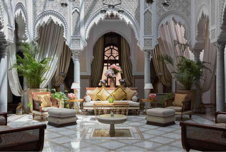 Intérieur d’un riad marocain de luxe à Marrakech, avec arches sculptées, motifs traditionnels, salon élégant aux coussins dorés, palmiers décoratifs et lumière naturelle mettant en valeur une esthétique raffinée et authentique.
