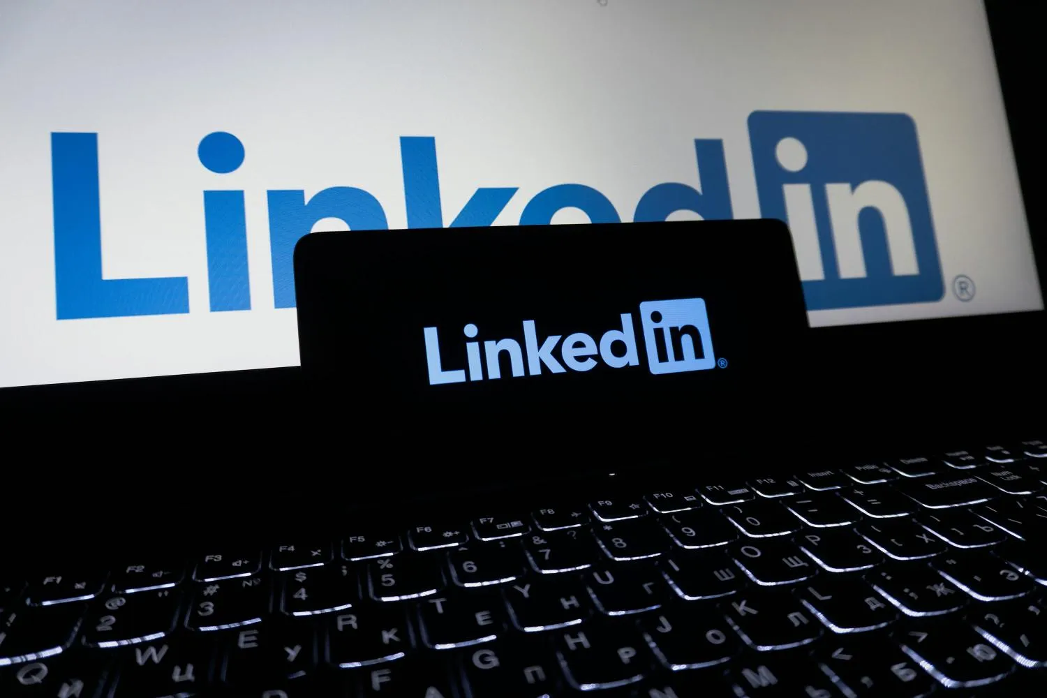 Logo LinkedIn affiché sur l’écran d’un ordinateur portable, avec un smartphone posé devant montrant également le logo LinkedIn sur fond sombre.