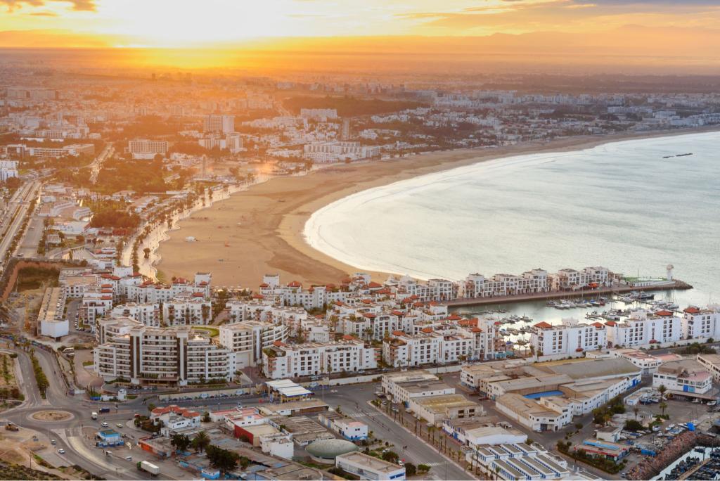 Vue aérienne haute définition de la baie d’Agadir au coucher du soleil, montrant la plage, la marina et le front de mer, symbole du branding touristique moderne et de la transformation visuelle des destinations balnéaires au Maroc.