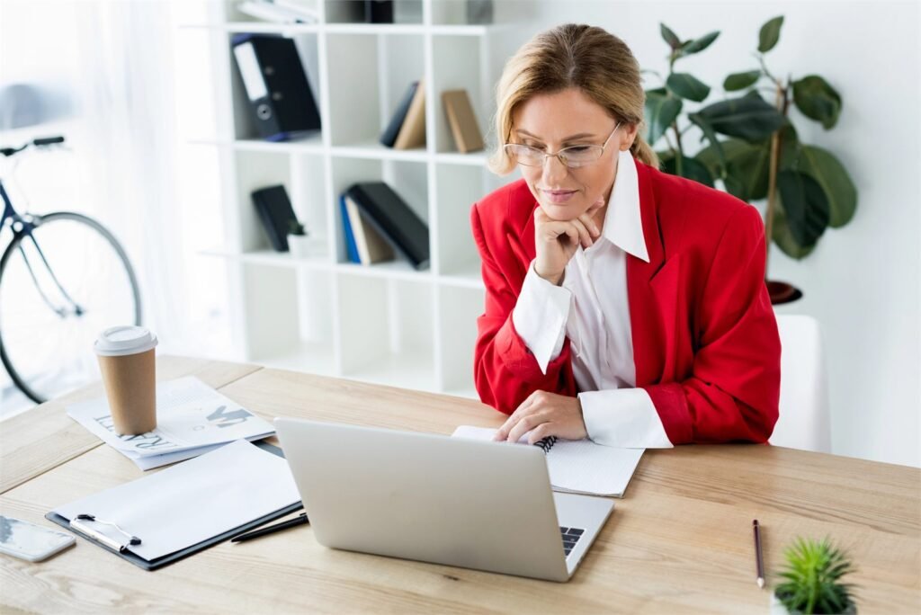 Business owner working at a laptop beside a notebook, clipboard and coffee in a bright office